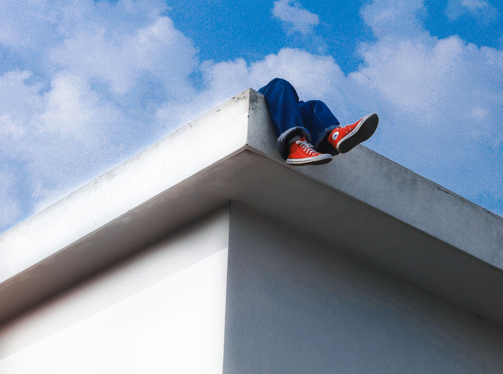 Looking up at pair of legs hanging over a roof amid a cloudy blue sky.