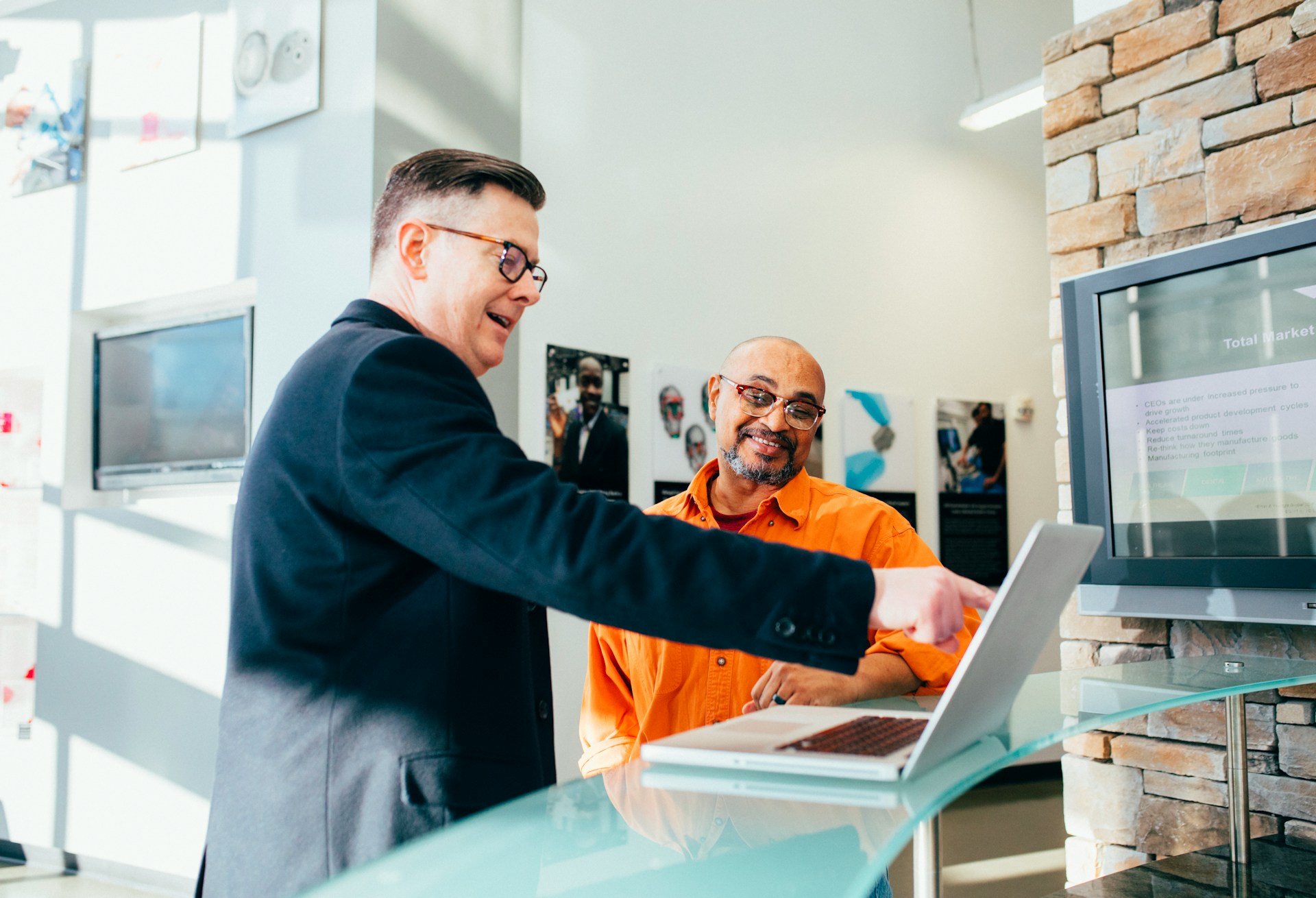 Two men near a laptop, one points to something on it and is saying something.